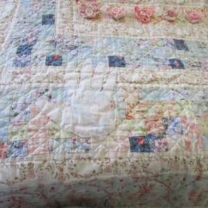 May include: A white fabric handprint appliqu&eacute;d on a patchwork quilt with floral patterns in shades of pink, blue, and white.