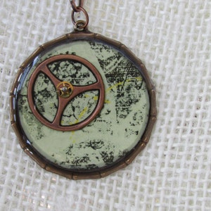 May include: A round pendant necklace with a copper-colored gear design and a green and white background. The pendant is made of resin and has a distressed look.