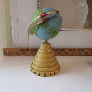 May include: A yellow and blue globe with a red and white rocket orbiting around it. The globe is on a yellow base with the word "FOSSIL" printed on it.