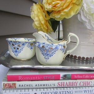 May include: A white creamer and sugar bowl set with blue and gold floral designs. The set is on a silver tray, which is on top of three books. The books are titled "Romantic Style", "Rachel Ashwell's Shabby Chic", and "Decorating with White".