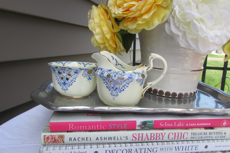 May include: A white creamer and sugar bowl set with blue and gold floral designs. The set is on a silver tray, which is on top of three books. The books are titled "Romantic Style", "Rachel Ashwell's Shabby Chic", and "Decorating with White".