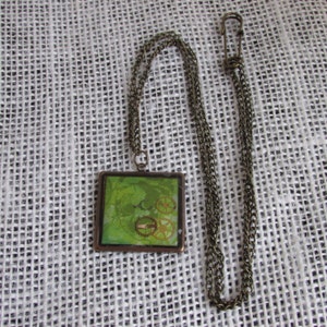 May include: A square green pendant with a bronze chain. The pendant features a design of gears and other mechanical elements.