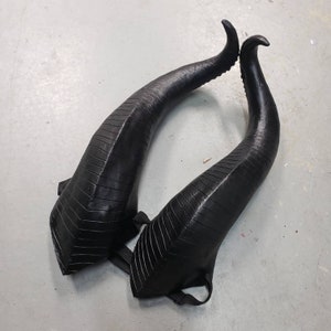 Puede incluir: Un par de cuernos negros, texturados y curvos. Los cuernos están hechos de un material flexible y tienen una punta ligeramente puntiaguda.