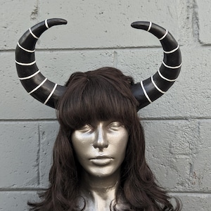 May include: A pair of black horns with white stripes, mounted on a mannequin head with dark brown hair. The horns curve upwards and outwards, creating a dramatic and fantasy-inspired look. The mannequin has a silver face and is set against a gray brick wall.