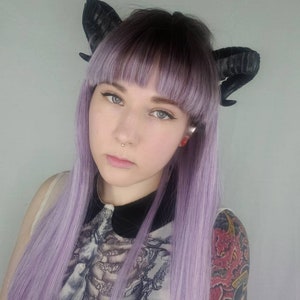 Puede incluir: Una persona con el pelo largo y lavanda y cuernos negros. Lleva una blusa transparente con estampado de esqueleto y un cuello negro. Tiene un piercing en la nariz y un tatuaje en el brazo.