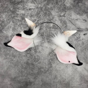 May include: A pair of white and black faux fur cat ears with pink inner ears, attached to a black headband.