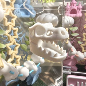 May include: A white 3D printed Tyrannosaurus rex skull figurine with an open jaw and visible teeth, positioned in front of a clear acrylic display case containing various pastel-colored decorative shapes including stars and clouds.
