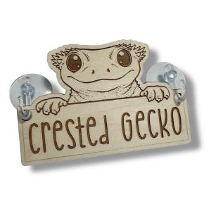 May include: A wooden sign with the text "Crested Gecko" and a cartoon gecko illustration. The sign is light brown and has two suction cups attached for hanging. The gecko has large eyes and is smiling.