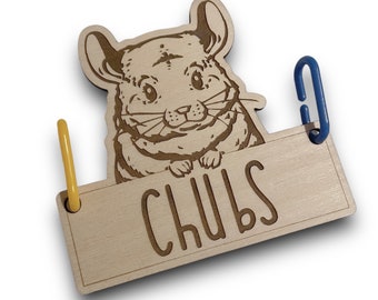 May include: Wooden sign with a chinchilla illustration and the name "CHUBS" engraved below. It has a yellow and blue metal clip. The chinchilla is light brown with etched details. A charming pet-themed item.