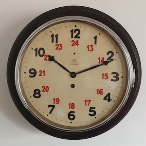 May include: A round wall clock with a dark brown frame and a cream-colored face. The dial features large black numerals from 1 to 12 and smaller red numerals from 13 to 24. It has black metal hands and the Junghans logo printed in the center.