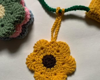 May include: A handmade crochet necklace featuring a yellow flower pendant with a brown center. The necklace has a green cord and a yellow loop. Additional crochet flower accents in green, pink, and blue are also visible.