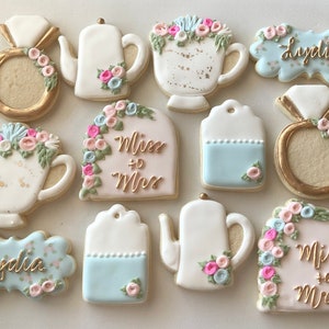 May include: Assortment of decorated sugar cookies in a bridal shower theme. Cookies include ring shapes, teacups, teapots, and cookies with the text "Lydia" and "Miss + Mrs". The cookies are decorated with pastel colors and floral accents.