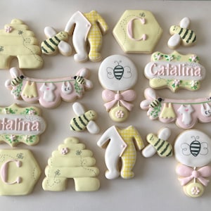 May include: A set of 14 yellow and pink decorated sugar cookies with bee and baby themes. The cookies feature a beehive, a baby onesie, a rattle, a banner with baby clothes, and the name "Catalina".