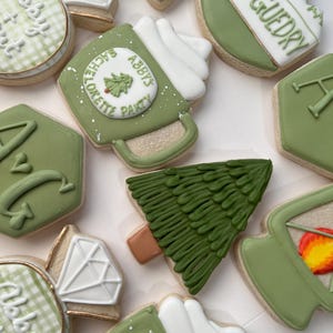 May include: An assortment of decorated sugar cookies in shades of green, white, and gold. The cookies feature various shapes, including a diamond, a mug, a Christmas tree, and a hexagon. Some cookies have text that reads "Abby's Bachelorette Party" and "Camp Guedry".