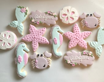 May include: Assortment of decorated sugar cookies in a sea life theme. Cookies include starfish, seahorses, mermaid tails, sand dollars, and crabs. Some cookies are personalized with the name "Adalynn". Colors include pink, white, and light blue.