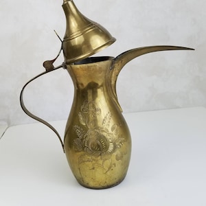 Può includere: Una caffettiera araba in ottone con un design floreale e un coperchio rimovibile. La caffettiera ha un beccuccio lungo e un manico.