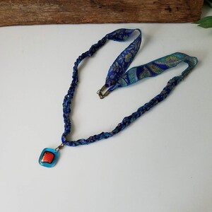 May include: A blue and turquoise braided fabric necklace with a square pendant featuring a red and gold design.