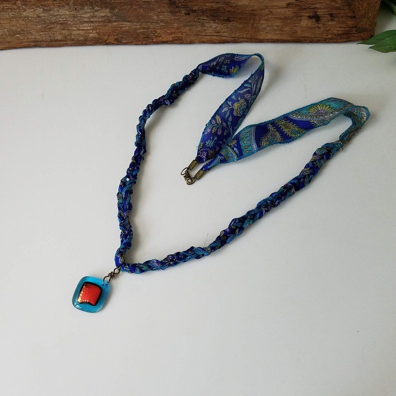 May include: A blue and turquoise braided fabric necklace with a square pendant featuring a red and gold design.
