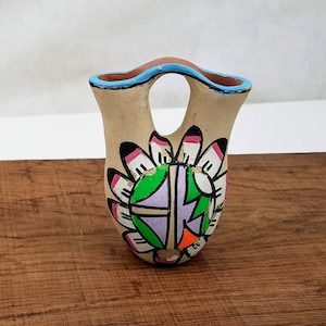 May include: A small, hand-painted ceramic vase with a light brown background and a colourful design. The design features a circle with green, purple and orange accents surrounded by white feathers with pink and black tips.