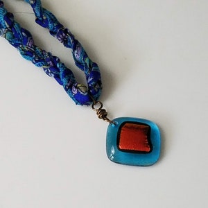 May include: A blue and white fabric necklace with a blue glass pendant featuring a red square inside.