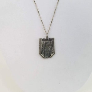 May include: A silver necklace with a pendant shaped like a shield. The shield has a design of a person holding a sword and a crown above their head. There are three smaller shields on the top of the shield.