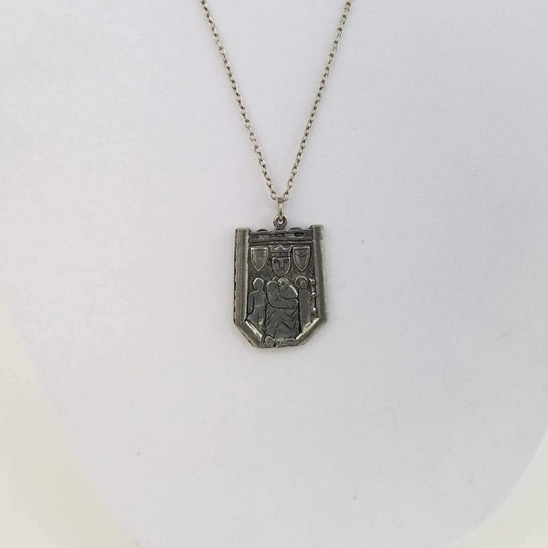 May include: A silver necklace with a pendant shaped like a shield. The shield has a design of a person holding a sword and a crown above their head. There are three smaller shields on the top of the shield.