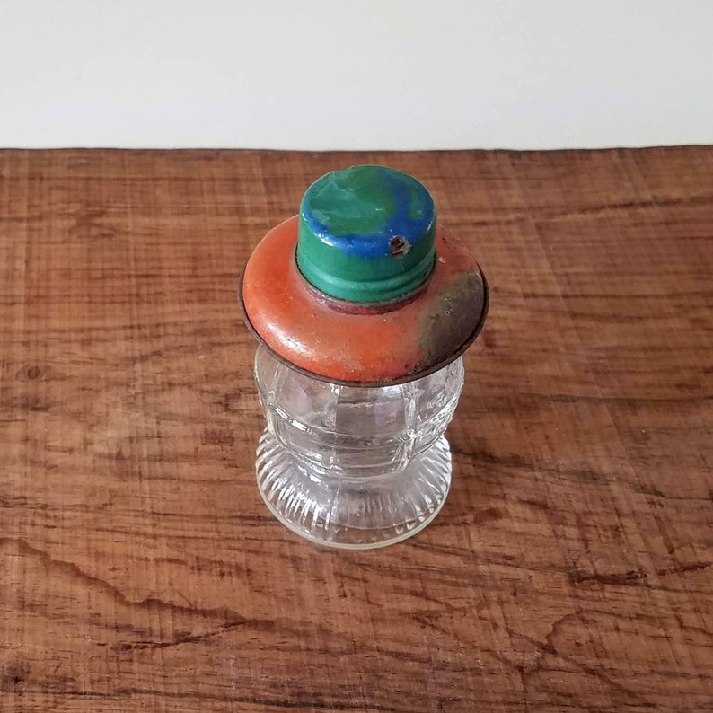 Puede incluir: Un tarro de vidrio vintage con una superficie texturizada y una tapa de metal. La tapa est&aacute; pintada de naranja, verde y azul.