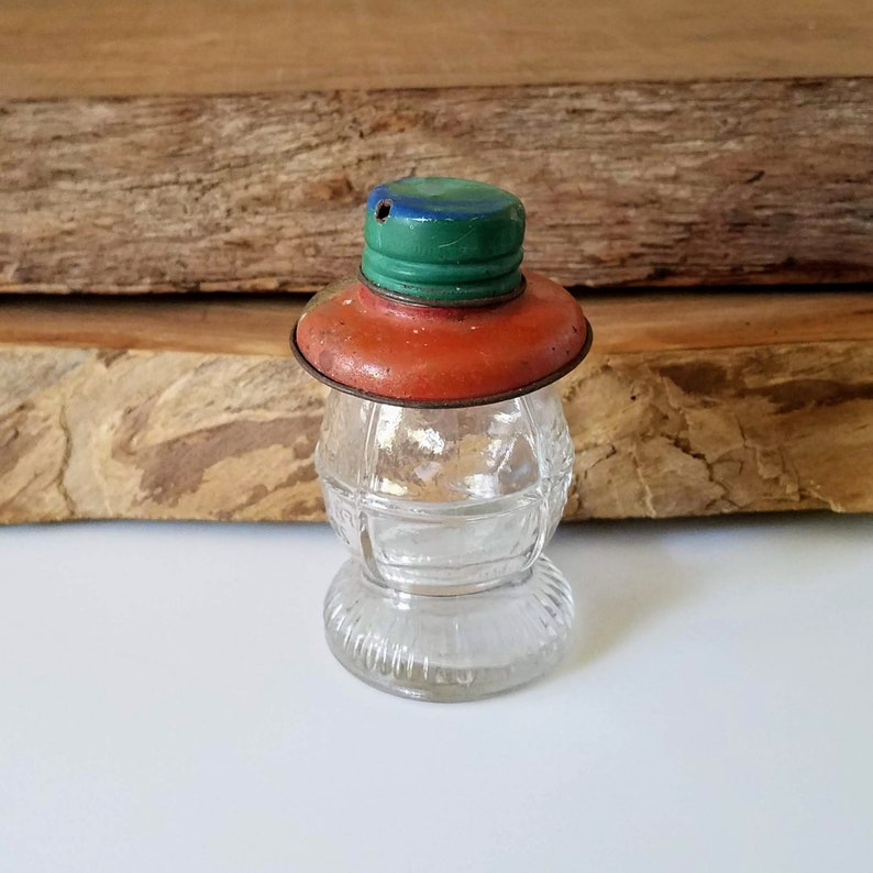 Puede incluir: Un tarro de vidrio vintage con una tapa de metal roja, verde y azul. El tarro tiene forma de linterna y tiene un cuerpo de vidrio transparente con un dise&ntilde;o texturizado.