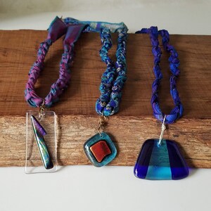 May include: Three necklaces with different colored glass pendants. The necklaces are made of braided fabric in shades of blue and purple. The pendants are a clear rectangle with a iridescent blue shard, a blue and orange square, and a blue triangle.