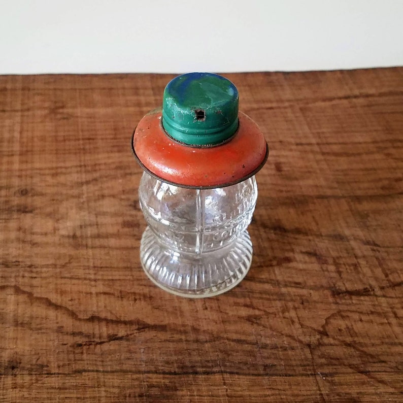 Puede incluir: Un frasco de vidrio transparente con una superficie texturizada y una tapa de metal roja y verde. La tapa tiene un peque&ntilde;o agujero en el centro.