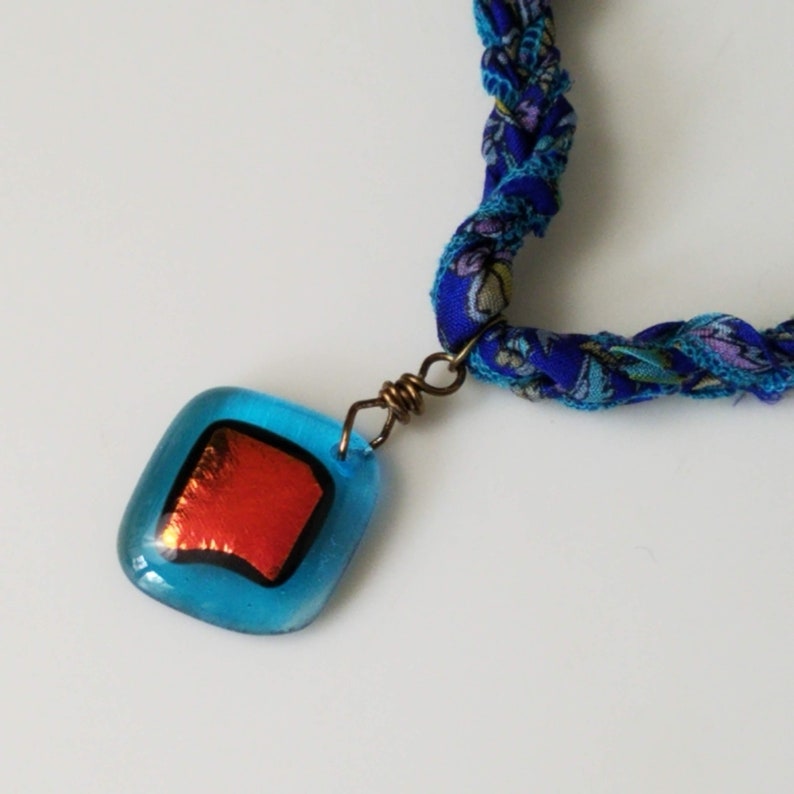 May include: A blue and red glass pendant hangs from a blue and purple braided fabric necklace. The pendant has a square shape with a red square inside.
