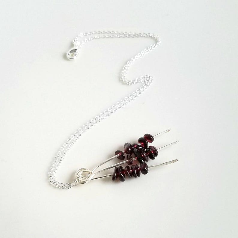 May include: A silver chain necklace with a pendant made of three silver wires with small, dark red garnet stones attached.