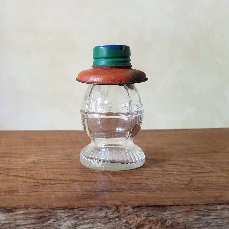 Puede incluir: Una botella de vidrio transparente con una superficie texturizada y una tapa de metal. La tapa est&aacute; pintada de naranja, verde y azul.