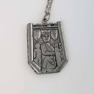 May include: Silver pendant necklace featuring a detailed relief of a crowned figure holding a child, surrounded by two other figures. The pendant is shaped like a shield with a decorative border.