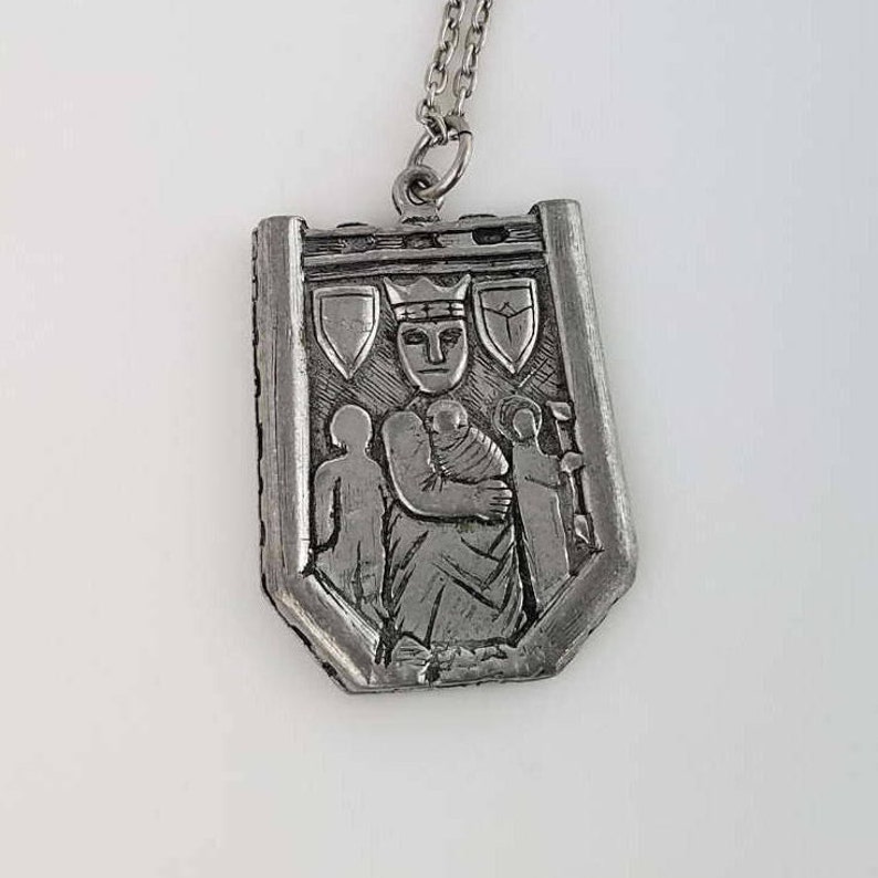 May include: Silver pendant necklace featuring a detailed relief of a crowned figure holding a child, surrounded by two other figures. The pendant is shaped like a shield with a decorative border.
