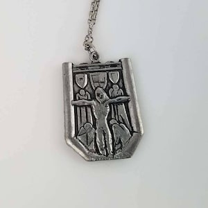 May include: A silver pendant necklace featuring a detailed relief of a person with outstretched arms, surrounded by figures in a gate-like structure.