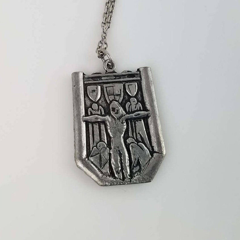 May include: A silver pendant necklace featuring a detailed relief of a person with outstretched arms, surrounded by figures in a gate-like structure.