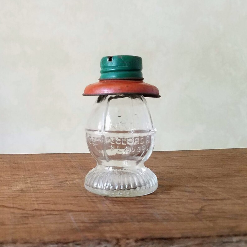 Puede incluir: Una botella de vidrio transparente con una superficie texturizada y una tapa verde y naranja. La botella tiene forma de bombilla y tiene el texto "Colors By" en el lateral.