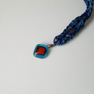 May include: A blue and white fabric necklace with a square blue glass pendant featuring a red square inside.