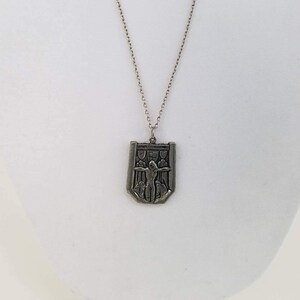 May include: A silver chain necklace with a pendant featuring a shield-shaped design with a raised image of a person with outstretched arms.