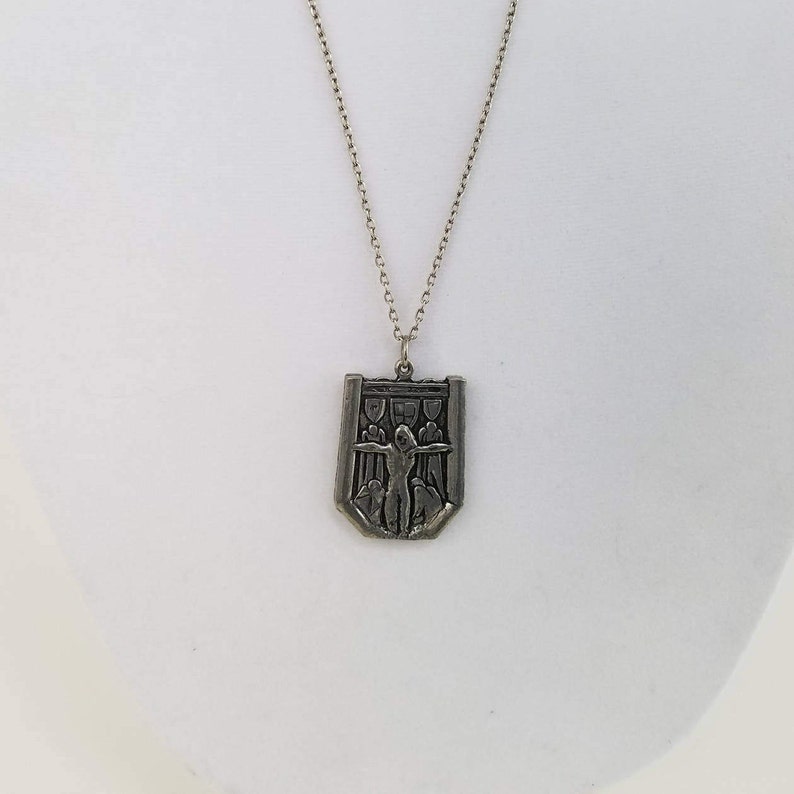 May include: A silver chain necklace with a pendant featuring a shield-shaped design with a raised image of a person with outstretched arms.