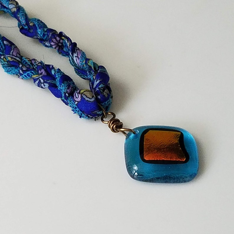 May include: A blue and purple fabric necklace with a square-shaped pendant. The pendant is made of glass and has a copper-colored square inside.