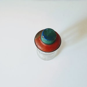 Puede incluir: Un tarro de vidrio vintage con una tapa roja, verde y azul. La tapa tiene un peque&ntilde;o agujero en el centro.