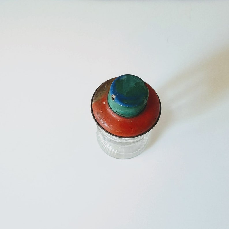 Puede incluir: Un tarro de vidrio vintage con una tapa roja, verde y azul. La tapa tiene un peque&ntilde;o agujero en el centro.