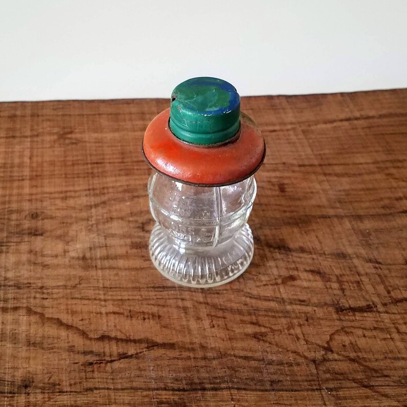 Puede incluir: Un frasco de vidrio transparente con una superficie texturizada y una tapa roja y verde. El frasco tiene un cuerpo redondo y una boca ancha.