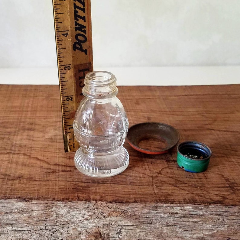 Puede incluir: Una botella de vidrio transparente con un dise&ntilde;o texturizado y una tapa de metal. La botella mide aproximadamente 7,5 cm de alto y tiene una forma redonda. La tapa es verde y tiene un anillo rojo alrededor.