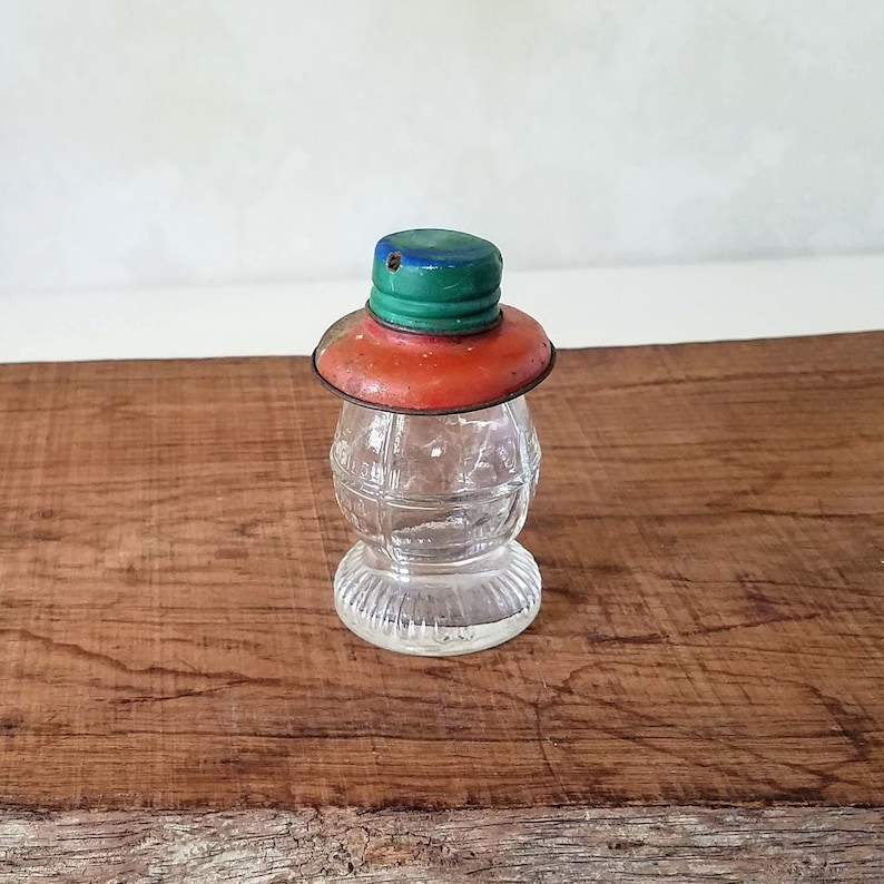 Puede incluir: Una botella de vidrio transparente con una tapa de metal roja y verde. La botella tiene forma de linterna y tiene una superficie texturizada.