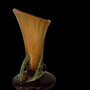 May include: A ceramic vase with a flared, trumpet-like shape. The vase is primarily a warm, earthy brown with a lighter, mottled interior. Green leaves and a small brown cone-like detail adorn the base, set against a black background.