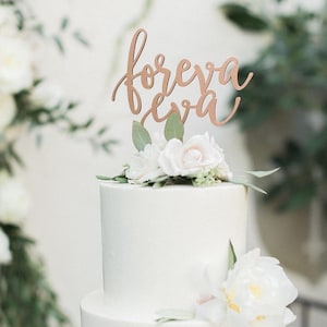 May include: A two-tiered white wedding cake with a wooden topper that says "foreva eva" in cursive script. The cake is decorated with white flowers and greenery.