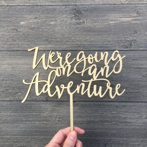 May include: Wooden cake topper with the text "We're going on an adventure" in a cursive font.